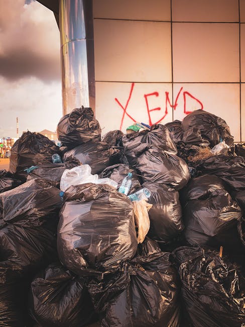 A large pile of black plastic rubbish bags, some of which are partially torn, piled against a modern building with a reflective metal surface and beige tiled wall. Several clear plastic water bottles are visible, some with caps, scattered among the bags. In the background, there is red graffiti text reading 'XEND' on the tiled wall. The scene is outdoors, seemingly on a paved or concrete surface, with an overcast sky and city skyline visible in the distance. The arrangement of bags suggests a waste collection or clearance situation, with the overall scene capturing an instance of unmanaged refuse likely requiring professional rubbish removal services to handle the accumulated waste efficiently and responsibly.