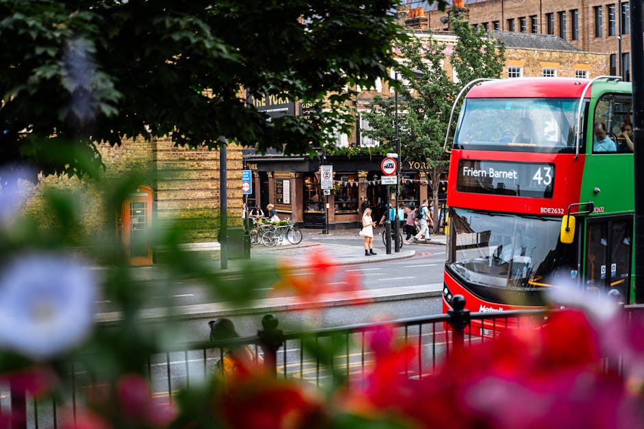 A double-decker bus with a red and green exterior is seen traveling along a street in an urban setting, with the front of the bus visible on the right side of the image displaying route information including the number 43 and the destination 'Friern Barnet.' In the foreground, colorful flowers and blurred green foliage partially obscure the scene, adding depth to the composition. Behind the bus, there is a crosswalk where pedestrians are walking across the street, some standing near bicycle racks or waiting to cross. The background features a variety of buildings, including a brick structure with multiple windows, and a shopfront with visible signage. The environment appears lively yet orderly, with natural light illuminating the scene, and the focus on the bus and pedestrians reflecting a typical busy day in an area that could benefit from professional rubbish removal services like those provided by houseclearancebarnet.co.uk for maintaining clean and tidy streets.