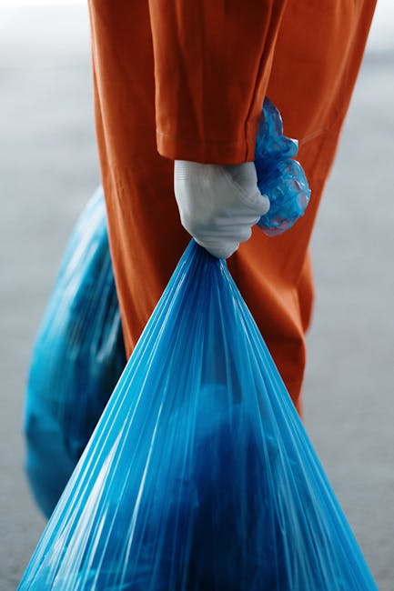 A close-up view of a person wearing orange protective overalls and a white glove, holding a large blue plastic garbage bag by its gathered top. The bag appears to be filled with waste material and has a glossy, semi-transparent surface with visible creases and folds. The individual is standing outdoors on a paved surface, with the background showing an out-of-focus urban or industrial environment. The scene suggests the person is engaged in waste collection or rubbish removal, possibly as part of an independent collection or on-site clearance service, exemplifying practices associated with private waste disposal services, aligned with the theme of rubbish removal. The lighting is natural, indicating daylight, and the focus remains on the hands, bag, and protective clothing, highlighting the professional approach to waste handling.