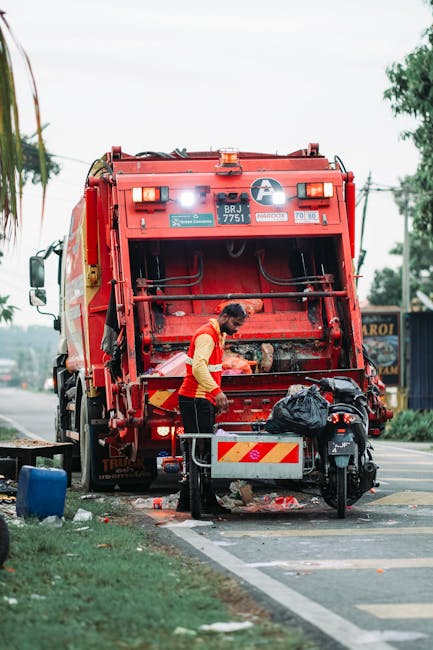 A worker in a yellow and red uniform is engaged in rubbish collection using a large red waste collection vehicle positioned on the roadside. The rear of the vehicle features a hydraulic compactor with metal components and safety markings, while the worker is manual handling waste materials, seen loading or unloading black plastic bags into the vehicle. The scene is set on a paved street with a petrol station or commercial building visible in the background, including signage, trees, and utility poles. The lighting indicates natural daylight, and the atmosphere appears to be a routine waste collection operation, consistent with private and independent disposal services that can offer alternative rubbish removal solutions outside of local authority provision. The presence of waste on the ground and bags in the worker's vicinity emphasizes the process of on-site clearance. The vehicle's structure and operational environment suggest an organised approach to rubbish removal, which companies like houseclearancebarnet.co.uk often support in handling various waste removal needs efficiently.