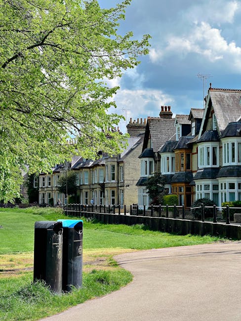 A row of traditional UK terraced houses with bay windows and slate-tiled roofs is visible in the background, set along a quiet residential street. In the foreground, on a grassy verge adjacent to a paved footpath, two black rubbish or recycling bins are positioned, with one slightly behind the other. A large, leafy tree with bright green foliage extends across the upper left corner of the image, partially shading the scene. The sky above is partly cloudy with patches of blue visible, adding natural daylight that illuminates the residential environment. The setting suggests an area where private waste collection or recycling services, such as those potentially offered by local providers like houseclearancebarnet.co.uk, might be used for rubbish removal, especially in contrast to standard local authority collection, emphasizing the context of independent waste handling in a suburban setting.