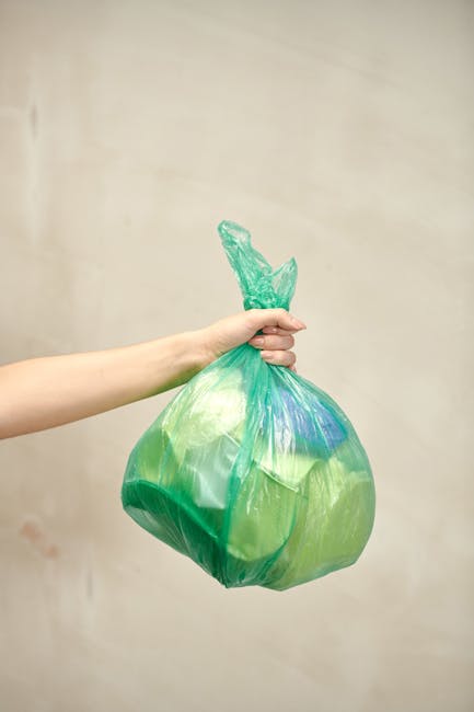 A person's hand is holding a semi-transparent green plastic rubbish bag filled with waste, knotted at the top and slightly bulging with contents. The bag appears to contain assorted refuse, possibly including paper and plastic items, and is held against a plain, light-colored wall background. The scene suggests an informal disposal or collection process, which may relate to private rubbish clearance services like those offered by houseclearancebarnet.co.uk, focused on domestic rubbish removal. The environmental context indicates a typical domestic setting, with the bag likely prepared for collection or disposal in an outdoor area such as a driveway or garden. The lighting is soft and even, emphasizing the textures and colours of the bag, with no other objects or environmental features visible in the frame.